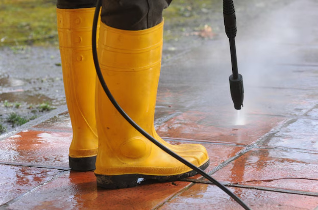 Person wearing yellow rubber boots with high-pressure water nozzle cleaning the dirt in the driveway